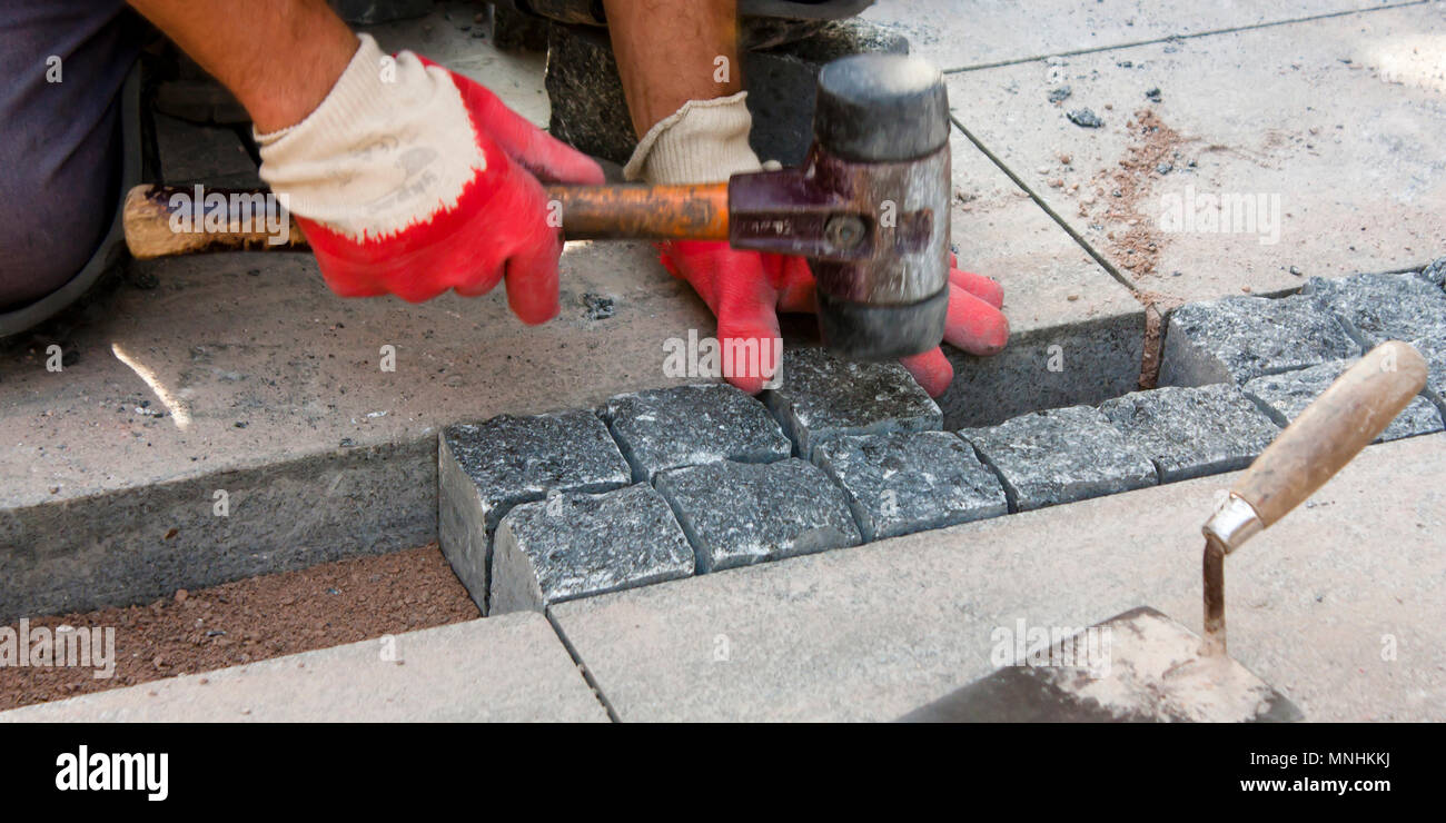 Le mani nei guanti di un lavoratore builder posa sul granito imp ciottoli sulla sabbia con un mazzuolo di gomma martello , e pavimentazione marciapiede di strada su strada con Foto Stock
