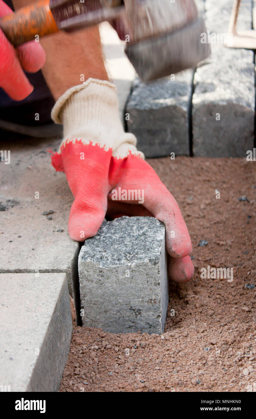 Le mani nei guanti di un lavoratore builder posa sul granito imp ciottoli sulla sabbia con un mazzuolo di gomma martello , e pavimentazione marciapiede di strada su strada con Foto Stock