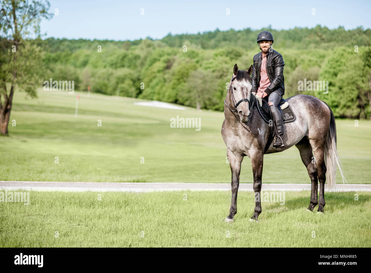 Uomo bello in giacca di pelle con casco protettivo in sella ad un cavallo sul prato verde Foto Stock