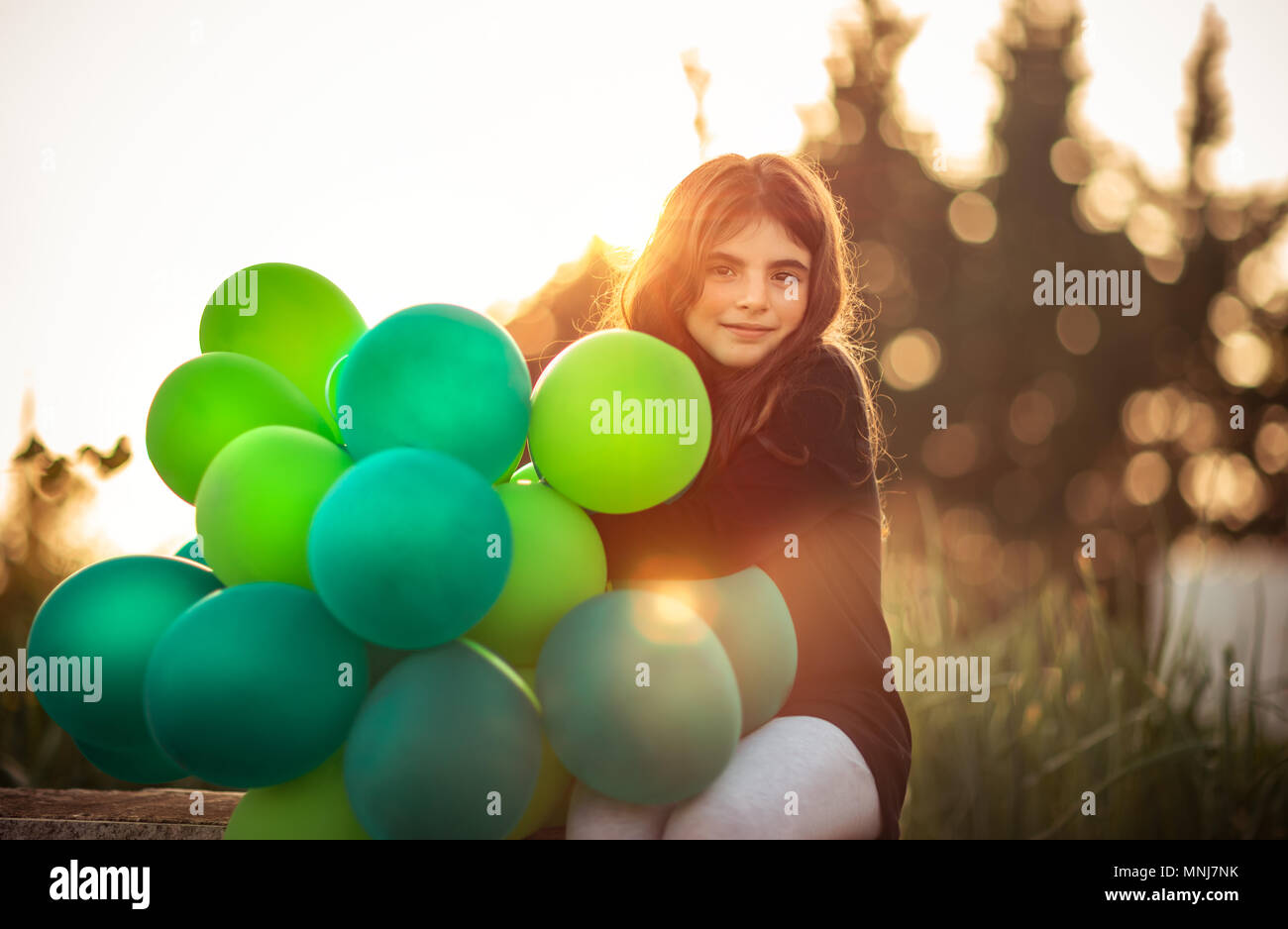 Ritratto di una incantevole piccola ragazza seduta nel parco con la pila di verde palloncini aria all'aperto in serata dolce della luce del sole, festeggia il compleanno, preparati Foto Stock