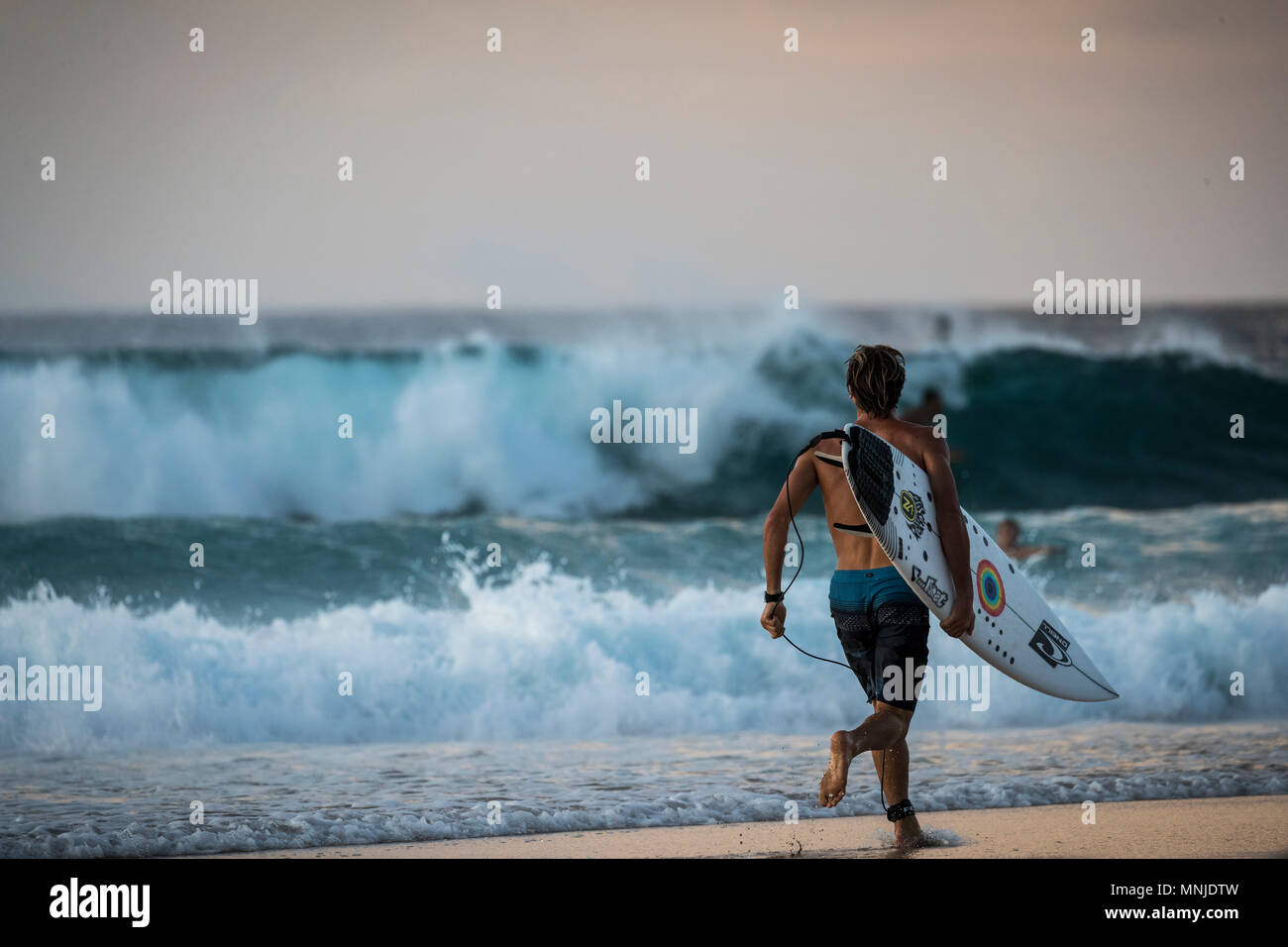 Vista posteriore del surfista maschio lungo la spiaggia al tramonto, Oahu, isole Hawaii, STATI UNITI D'AMERICA Foto Stock