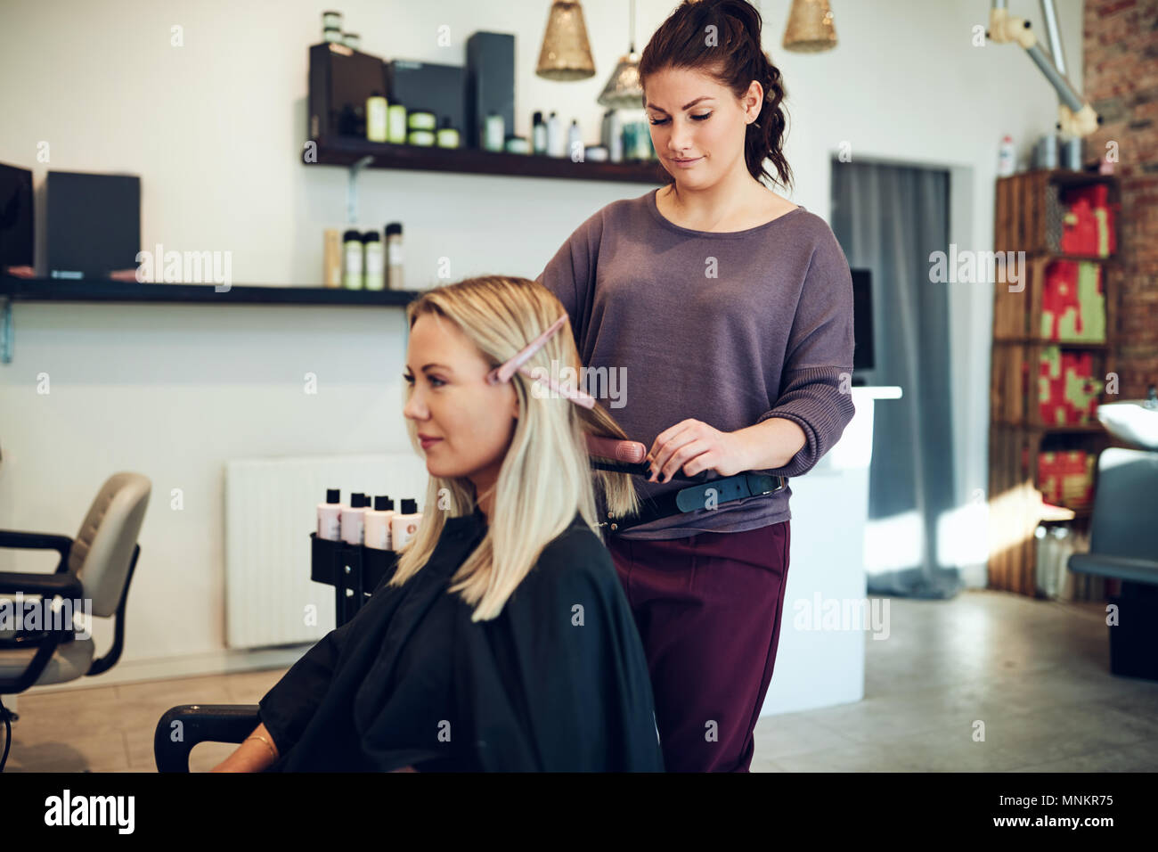 Bionda e giovane donna seduta su una sedia in un salone di bellezza con i suoi capelli raddrizzato dal suo parrucchiere Foto Stock
