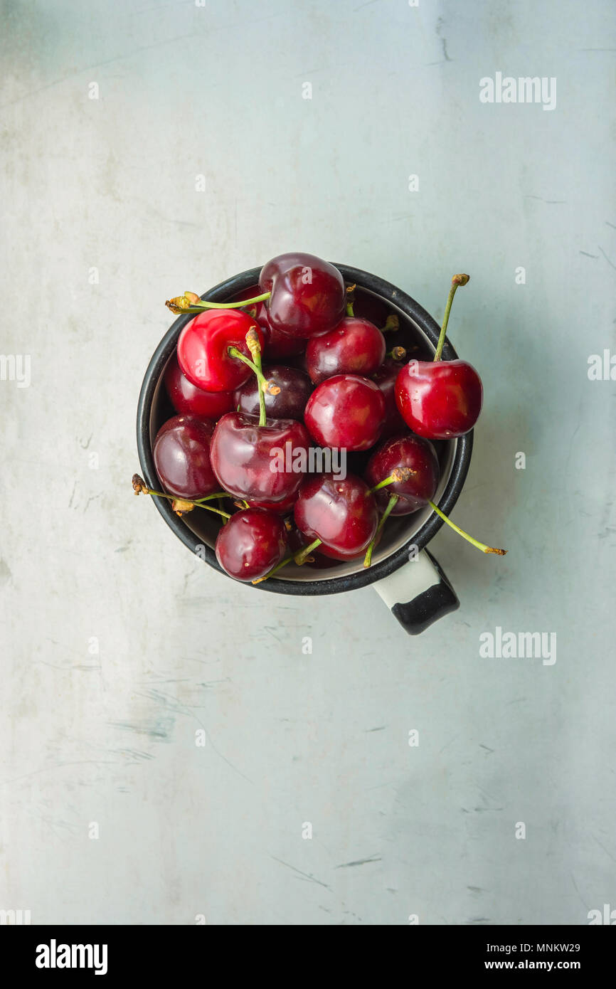 Organico mature appena raccolte le ciliegie dolci in smalto Vintage Mug in metallo grigio pietra sullo sfondo di calcestruzzo dalla finestra. Tavolo da giardino. Lay piatto. Estate Har Foto Stock