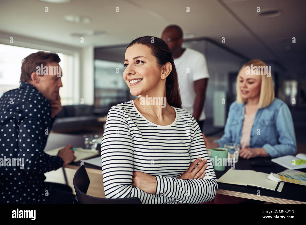 Giovane imprenditrice guardando sopra la sua spalla e sorridere mentre seduto con i colleghi a una tabella in un ufficio Foto Stock