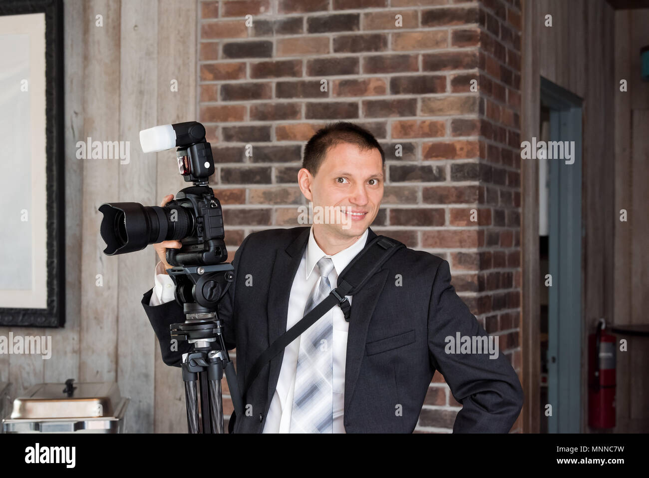 Un giovane sorridenti fotografo di matrimoni in tuta e cravatta in piedi con fotocamera flash esterni, treppiede all'interno sala ristorante edificio Foto Stock