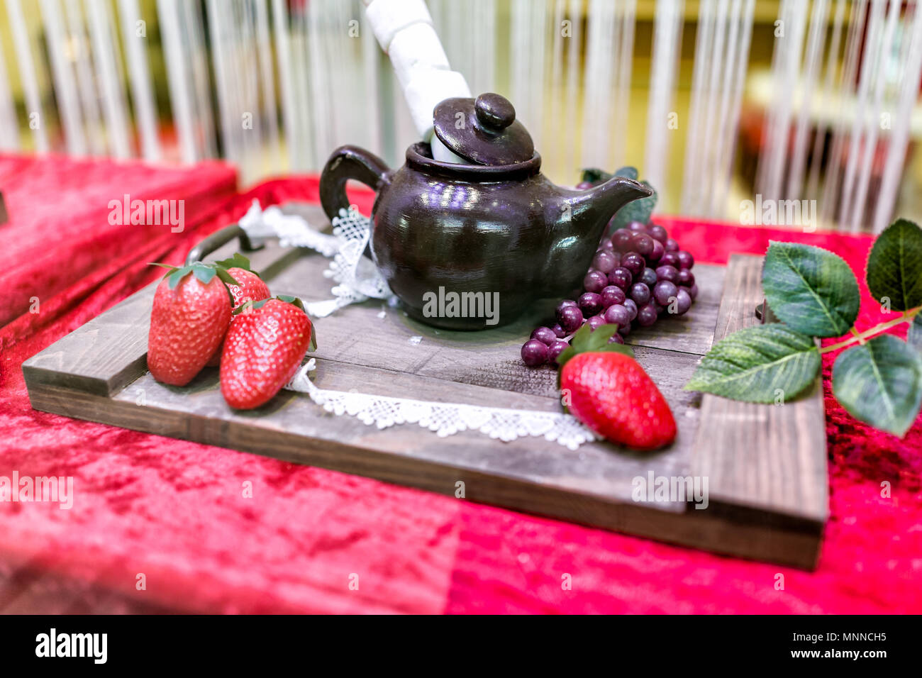 Visualizzazione di cioccolato commestibile teiera con marshmallows, fragole, sul tagliere di legno decorazione con foglie verdi e uve frutto, velluto rosso Foto Stock
