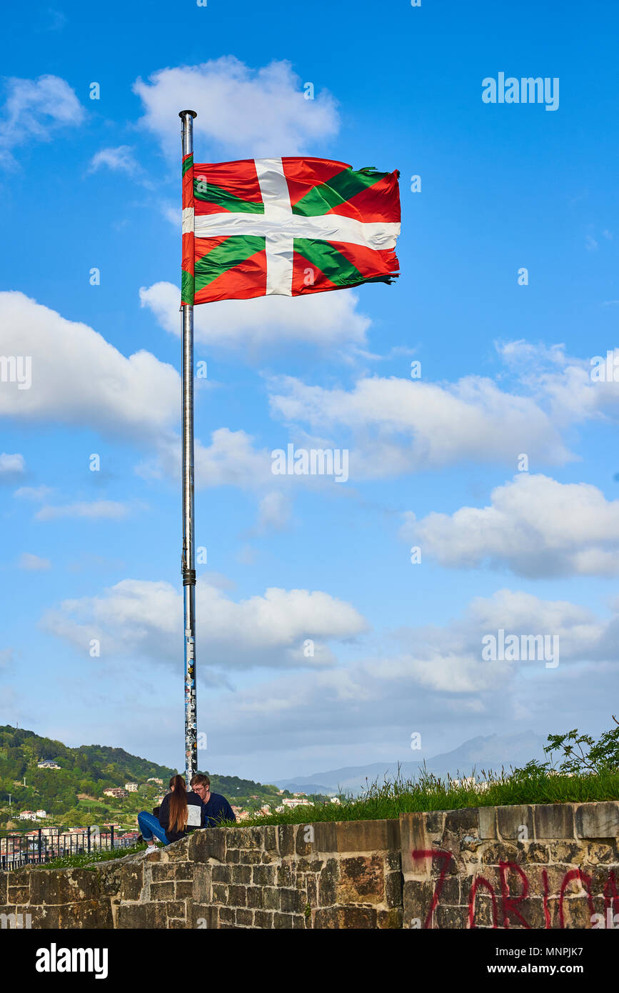 Due giovani seduti sotto il montante di una Ikurrina, Paesi Baschi bandiera sventola su un cielo blu nel Monte Urgull di San Sebastian. Spagna. Foto Stock