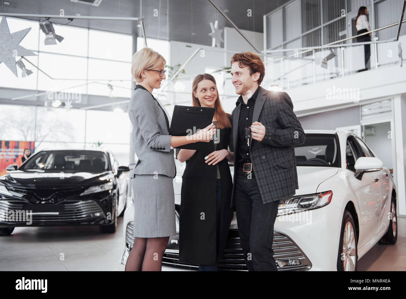 Di acquistare la loro prima vettura insieme. Angolo di Alta Vista del giovane venditore auto in piedi presso la concessionaria dicendo circa le caratteristiche della vettura per i clienti Foto Stock