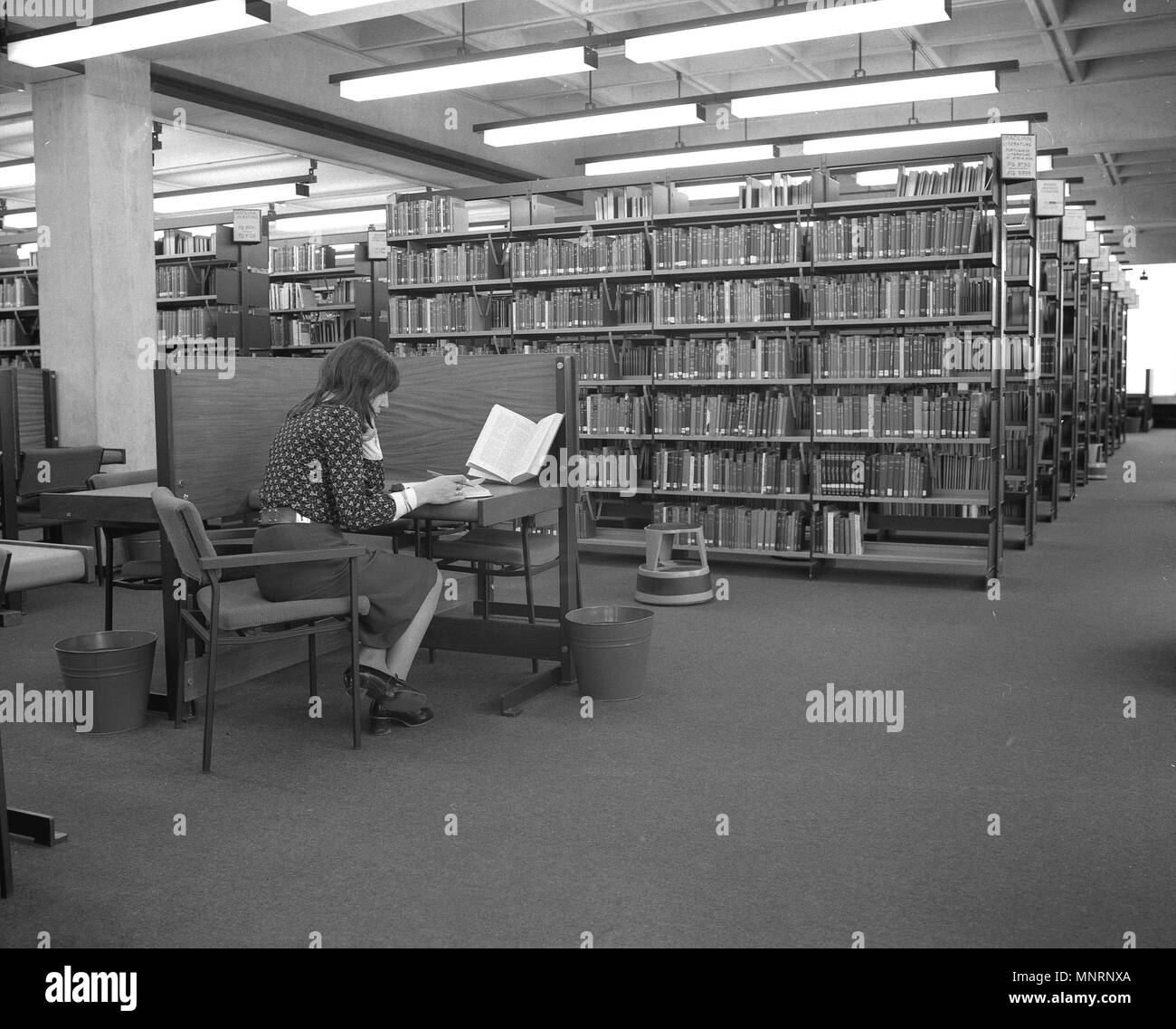 Anni sessanta, storico, una giovane donna da solo in una grande e moderna biblioteca seduti ad un piccolo tavolo con schermo, studiando un libro, Inghilterra, Regno Unito. Foto Stock