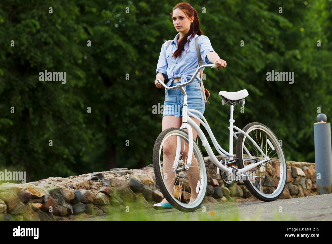 Ragazza in pantaloncini blu e una camicia in denim, passeggiate su asfalto con bicicletta Bianchi serata estiva in città, sullo sfondo di verdi alberi, muri di pietra Foto Stock