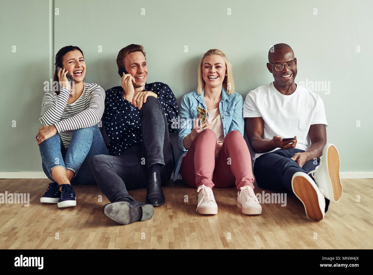 Gruppo di diversi giovani imprenditori seduti insieme su un ufficio pavimento durante breaktime utilizzando i loro telefoni cellulari Foto Stock