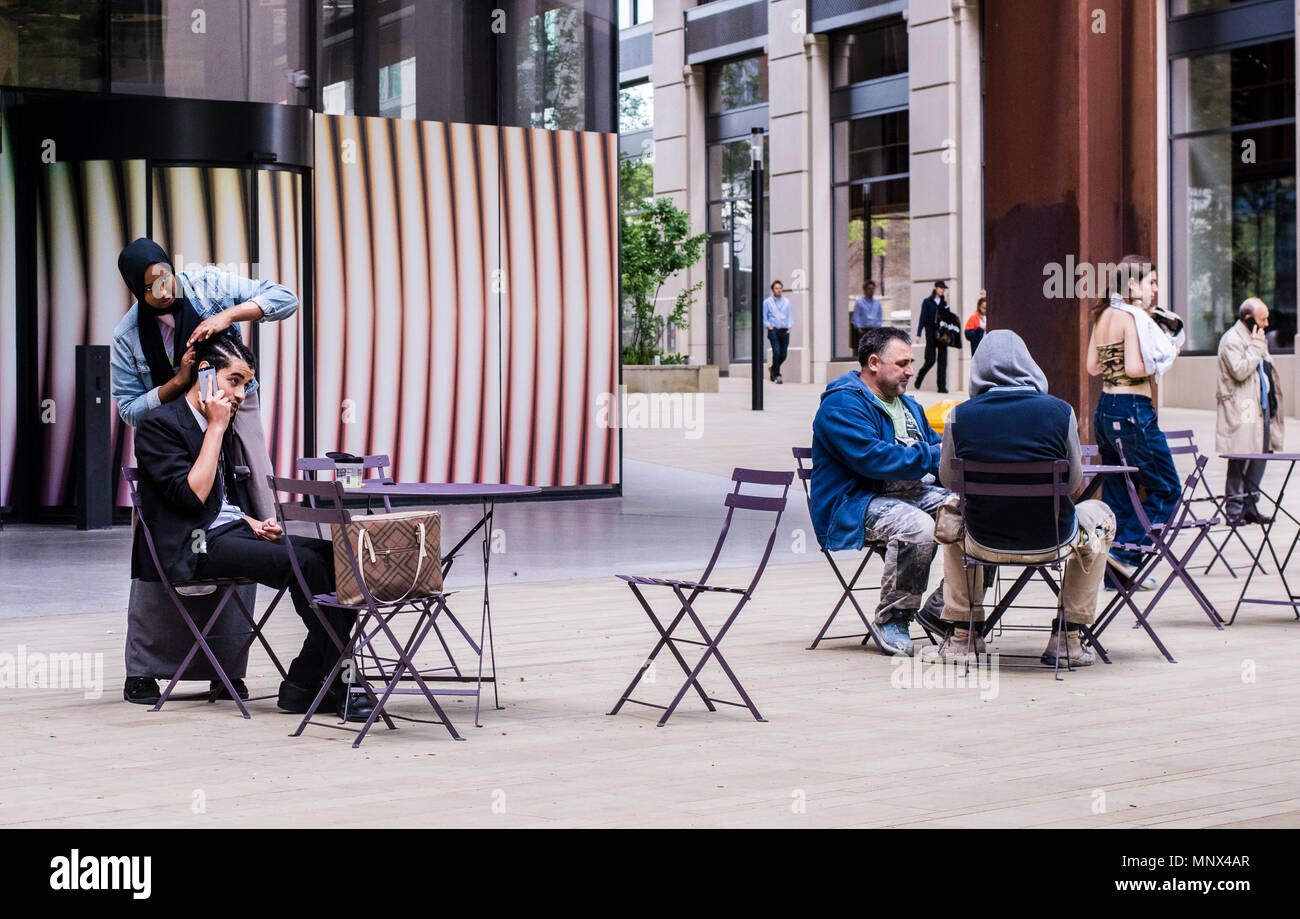 Giovane uomo seduto a un tavolo bar parlando sul suo telefono cellulare, mentre una ragazza lavora sui suoi dreadlocks, Kings Cross, London, England, Regno Unito Foto Stock