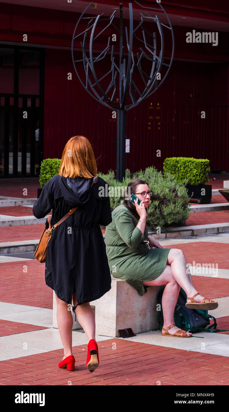 Donna con i capelli rossi, indossando il colore rosso brillante scarpe, a piedi dalla fotocamera, un'altra donna nelle vicinanze, seduta, parlando al telefono, London, England, Regno Unito Foto Stock