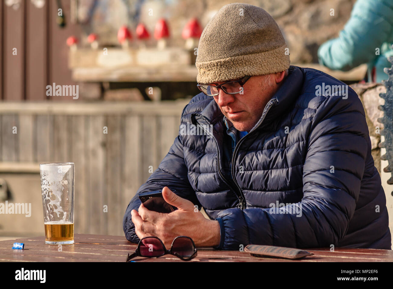 Uomo in giacca calda e hat sat al di fuori di un pub con una quasi finito una pinta di birra e utilizzando un telefono cellulare. Teignmouth, Devon. Feb 2018. Foto Stock