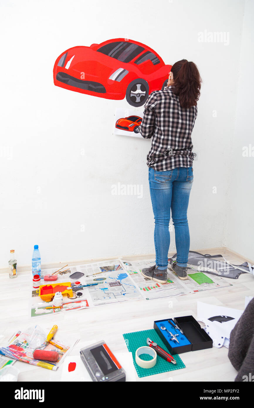 Close-up di una giovane donna artista e la madre del ragazzo per il disegno di un bambino su un muro bianco un bel rosso auto in una luce stanza dei bambini, sul pavimento giace Foto Stock