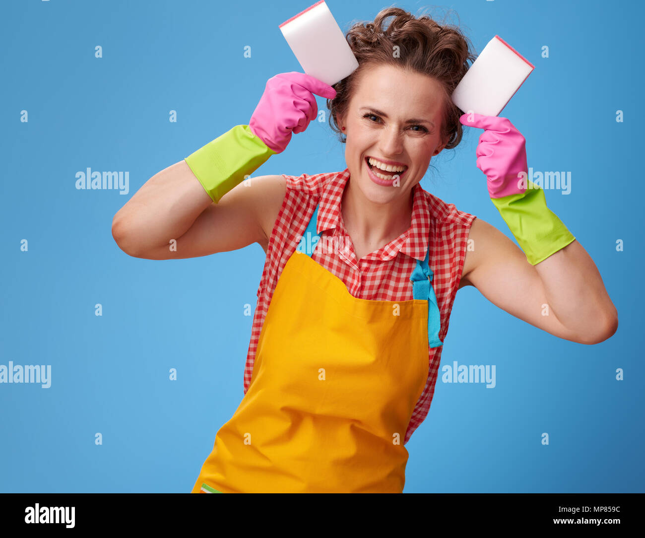 Grande tempo di pulizia. Felice giovane casalinga con guanti di gomma rendendo le orecchie con spugne cucina isolato su blu Foto Stock