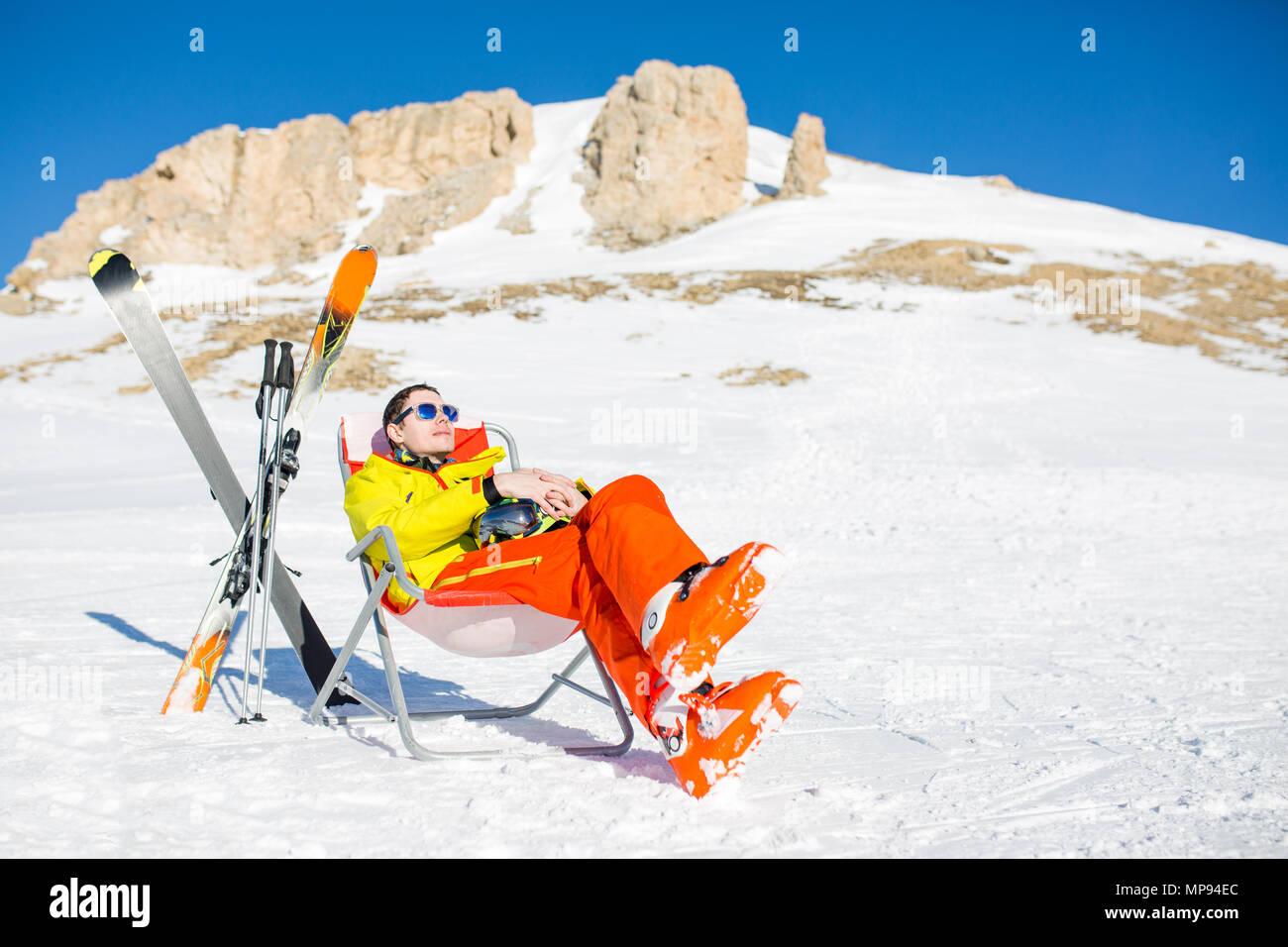 Immagine di sport uomo seduto sulla sedia accanto a sci e bastoni sullo sfondo di montagne innevate Foto Stock