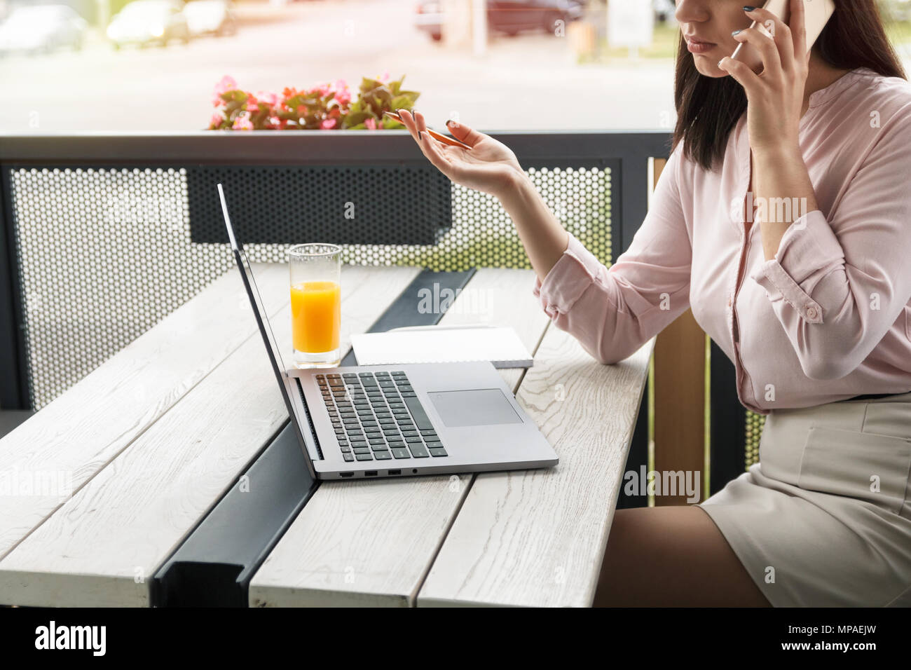 Giovane donna di affari che parla al telefono e gesticulating tenendo una matita, laptop e succo di frutta sulla scrivania. Foto Stock