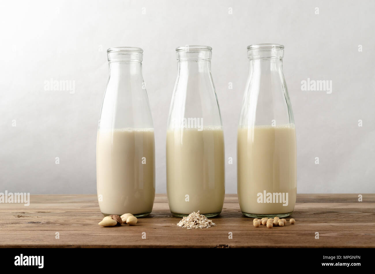 Fila di tre bottiglie di vetro contenenti senza latte e derivati del latte bevande sostitutive. Ingredienti chiave di mandorle; avena e i semi di soia in primo piano su legno planked Foto Stock