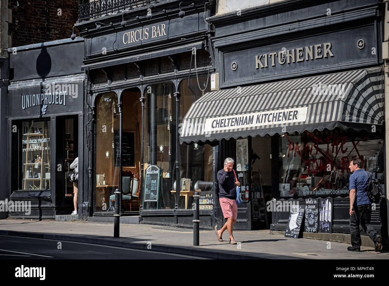 I pedoni a camminare lungo la strada al di fuori di nero fronteggiata negozi e bar sul lungomare vicino a Montpellier, Cheltenham, Gloucestershire. Foto Stock