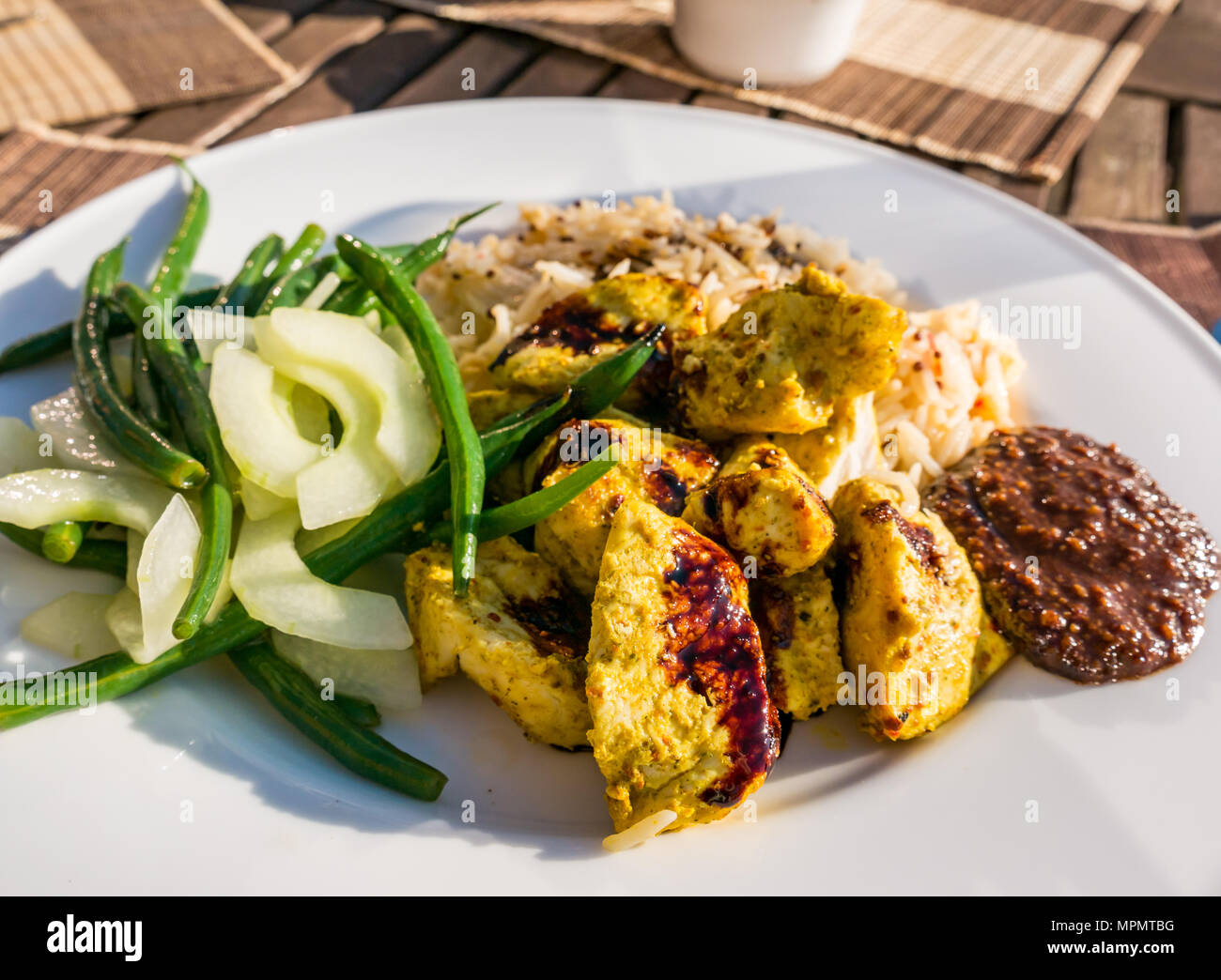 Pasto indonesiana servita su bianco a cena la piastra sul patio tabella nella luce del sole. Pollo satay, salsa di arachidi, kecap masin salsa, riso, fagioli verdi & cetriolo Foto Stock