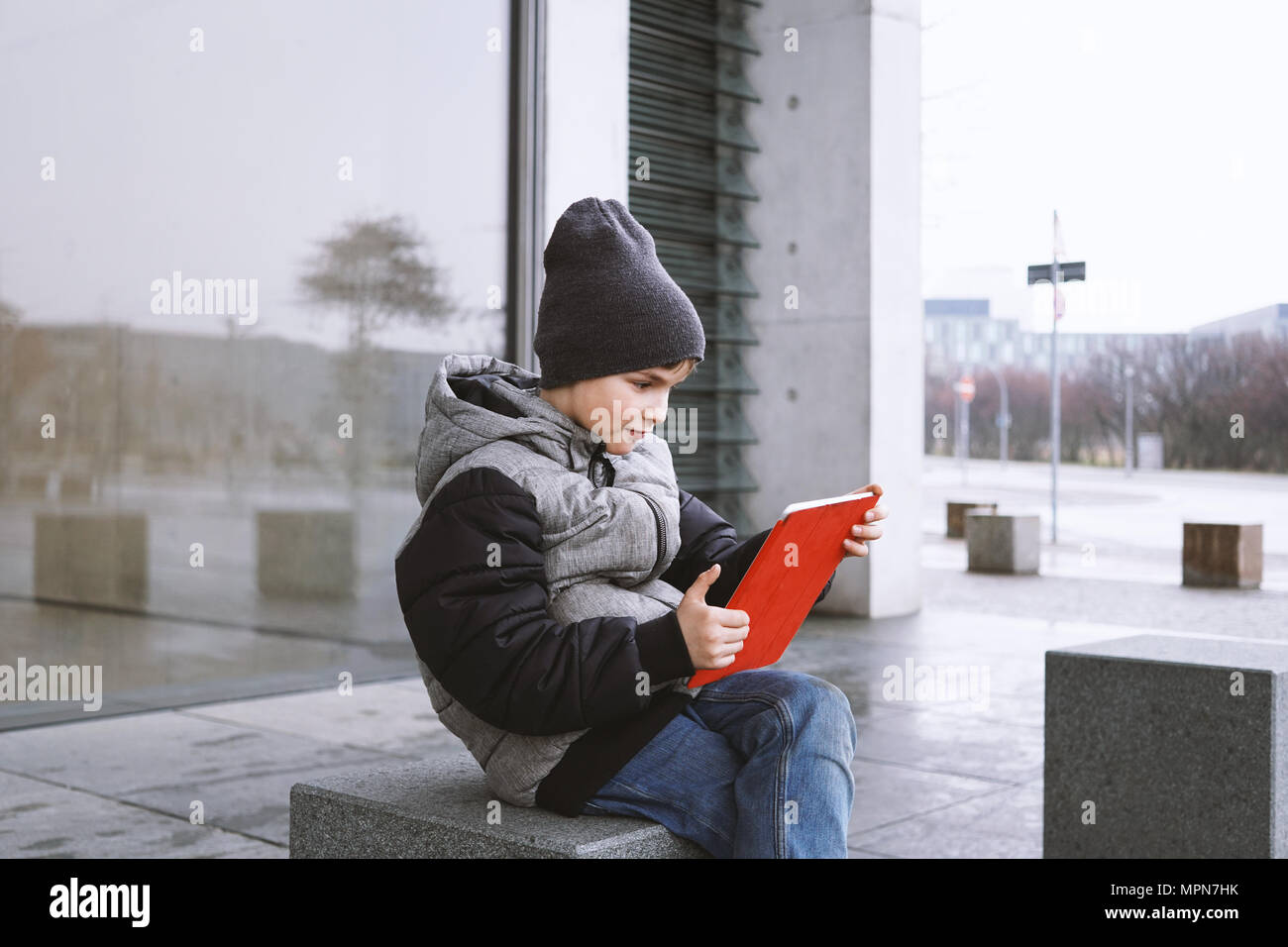 7 anno vecchio ragazzo giocando online gioco sul computer tavoletta mentre seduto fuori da solo sulla strada di città in inverno Foto Stock