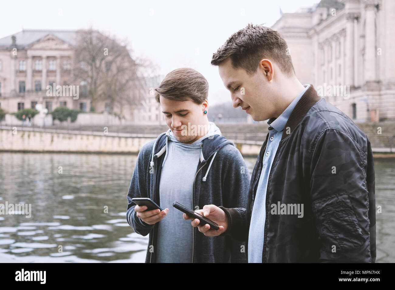 Due antisociale di telefono cellulare di tossicodipendenti adolescenti di sesso maschile in piedi insieme utilizzando lo smartphone, concetto tecnologico, urban posizione sulla riva del fiume a Berlino Germania Foto Stock