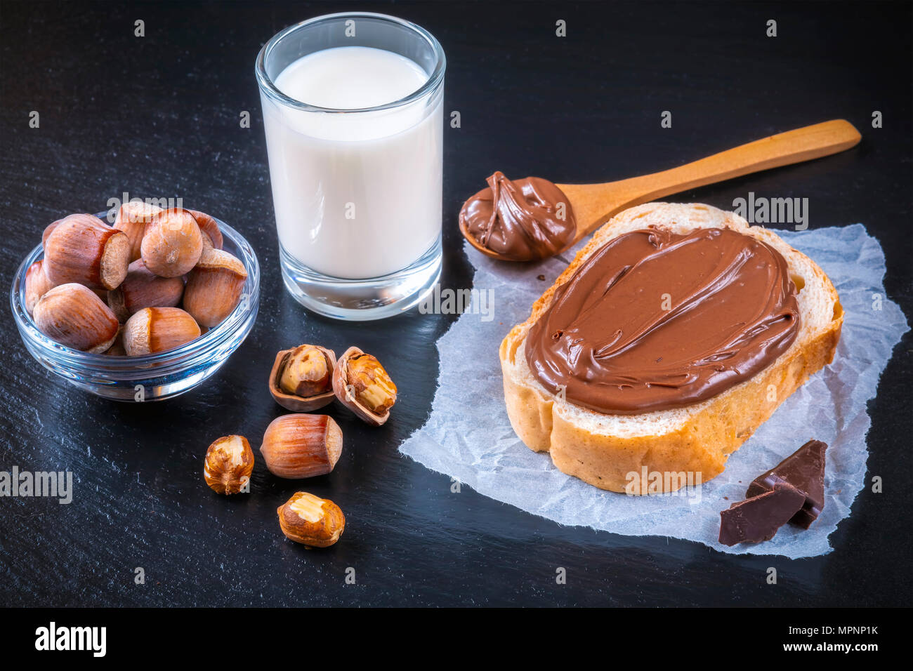 Fetta di pane con il diffondersi di crema di cioccolato e nocciole sulla tavola nera. Serve una deliziosa colazione concetto. Focus su nocciola nella ciotola. Foto Stock