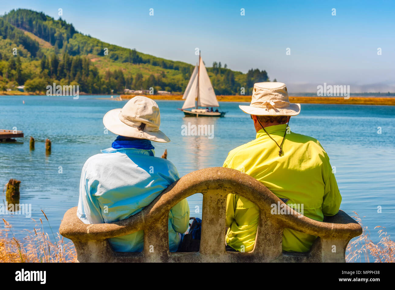 Wheeler, Oregon,USA = Agosto 15, 2012: una coppia seduta su una panchina lungo le rive di Tillamook Bay come una barca a vela vele passato in Wheeler, Oregon Foto Stock