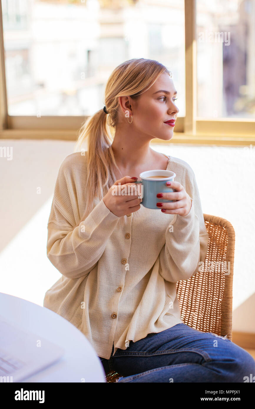 Donna bionda con tazza di caffè guardando fuori della finestra Foto Stock