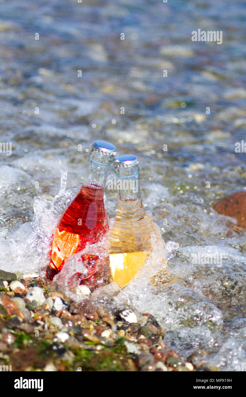 Due bottiglie sono in piedi in acqua sulla spiaggia in estate. Rosso e giallo di bevande. Foto Stock