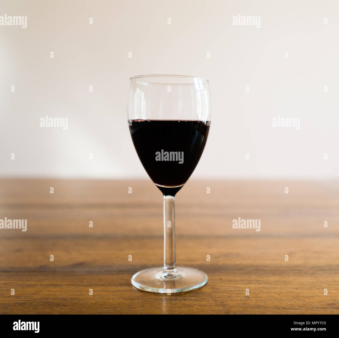 Un bicchiere mezzo pieno di vino rosso scuro in piedi sulla tavola di legno contro il muro bianco sfondo sfocato. Foto Stock
