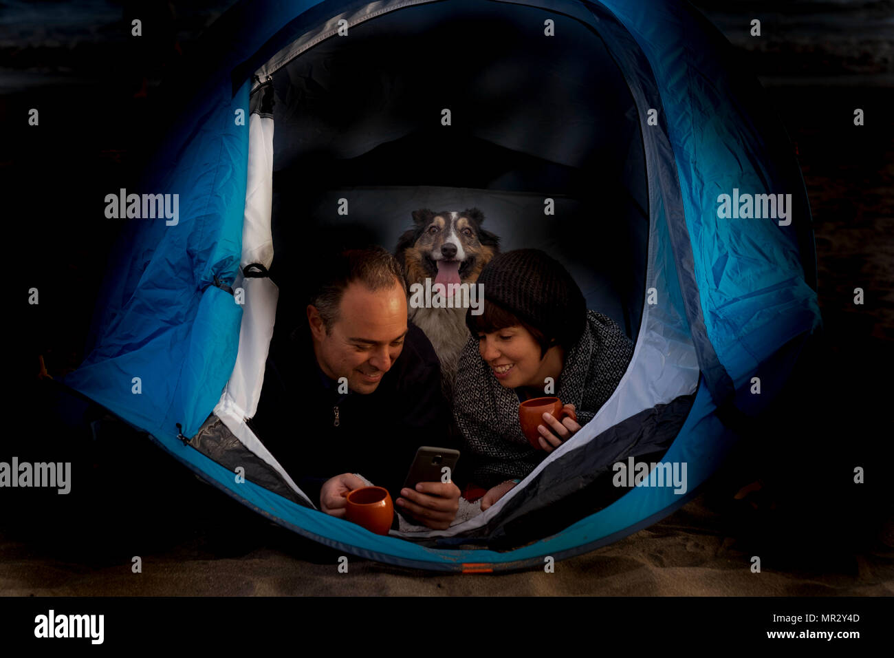 Campeggio vacanza alternativa lifestyle con coppia di mezza età con Border Collie cane dentro una tenda di notte. Entrambi bere il tè o il caffè e controllare la m Foto Stock