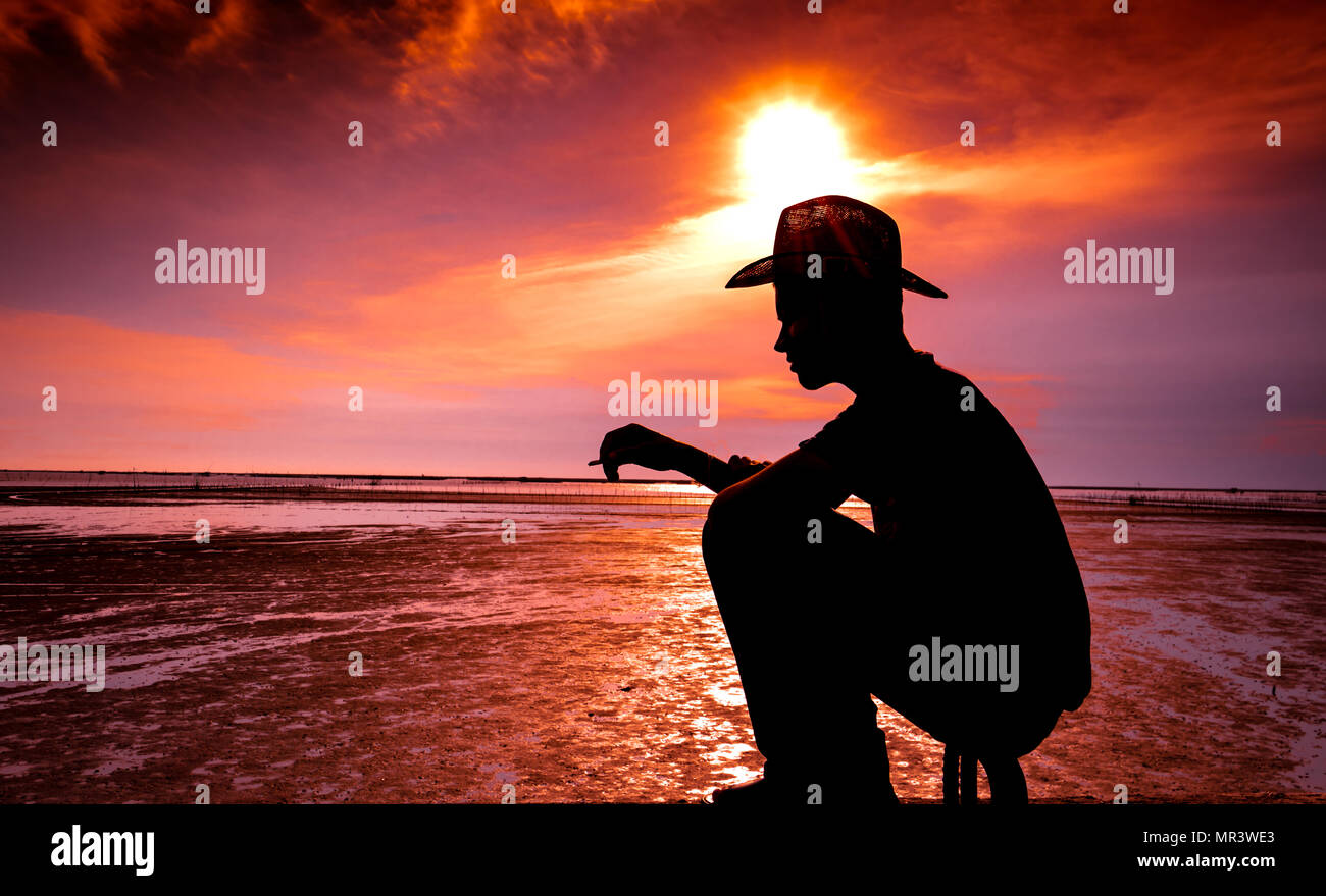 Silhouette di un giovane uomo seduto sulla spiaggia in riva al mare e il fumo nella sera al tramonto sulla sua vacanza. La Lonely Man provate a rilassarvi e thi Foto Stock