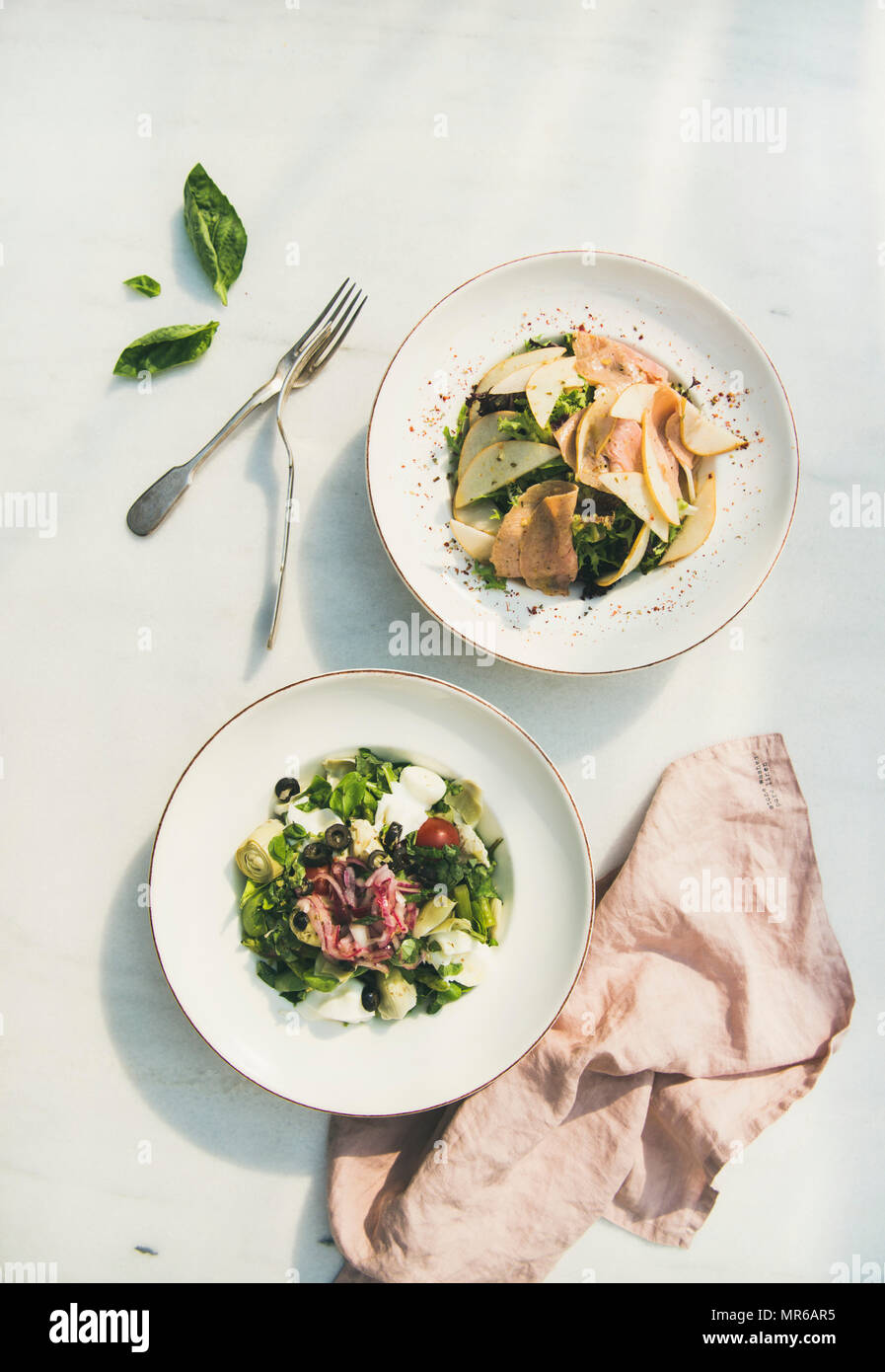 Flat-lay di fresco e verde insalata estiva con carciofi, olive e formaggio morbido e cipolla rossa e posate in bianco su lastre di marmo chiaro tabella backgroun Foto Stock