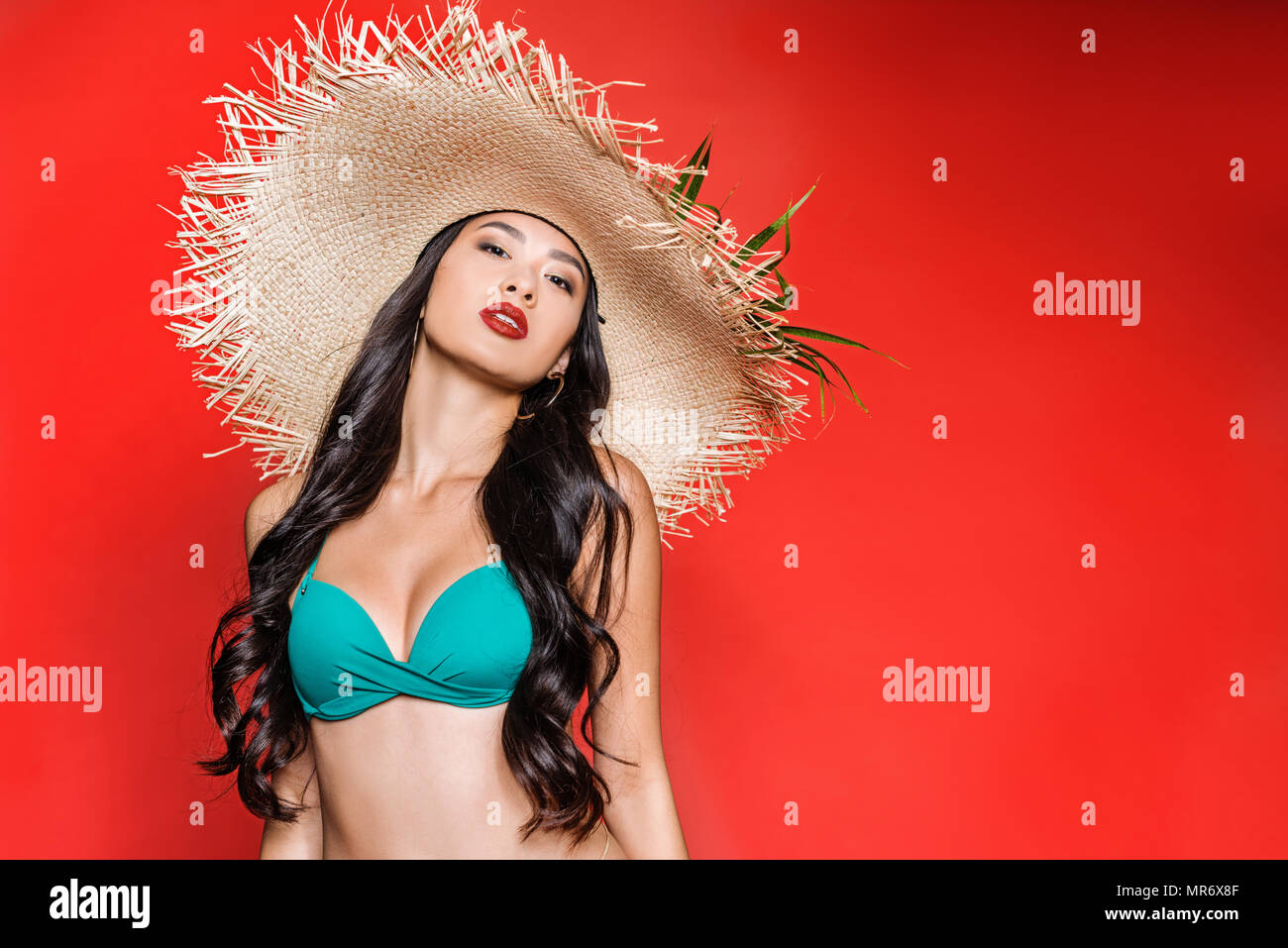 A mezza lunghezza shot di bella donna asiatica in costume da bagno luminoso e cappello di paglia che posano su rosso Foto Stock