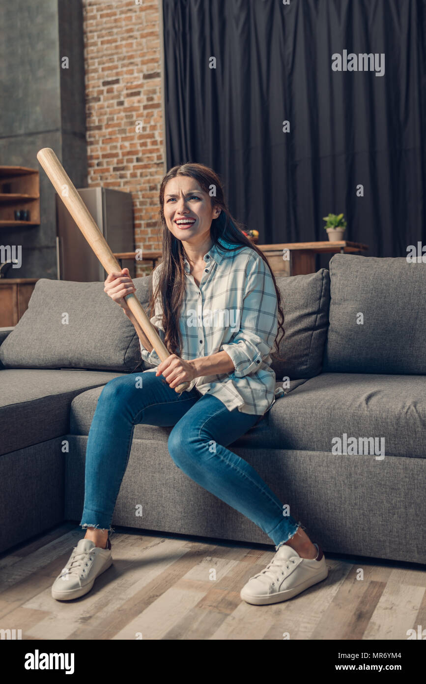 Giovane donna in possesso di una mazza da baseball seduto sul divano nel soggiorno e la visione di un gioco Foto Stock