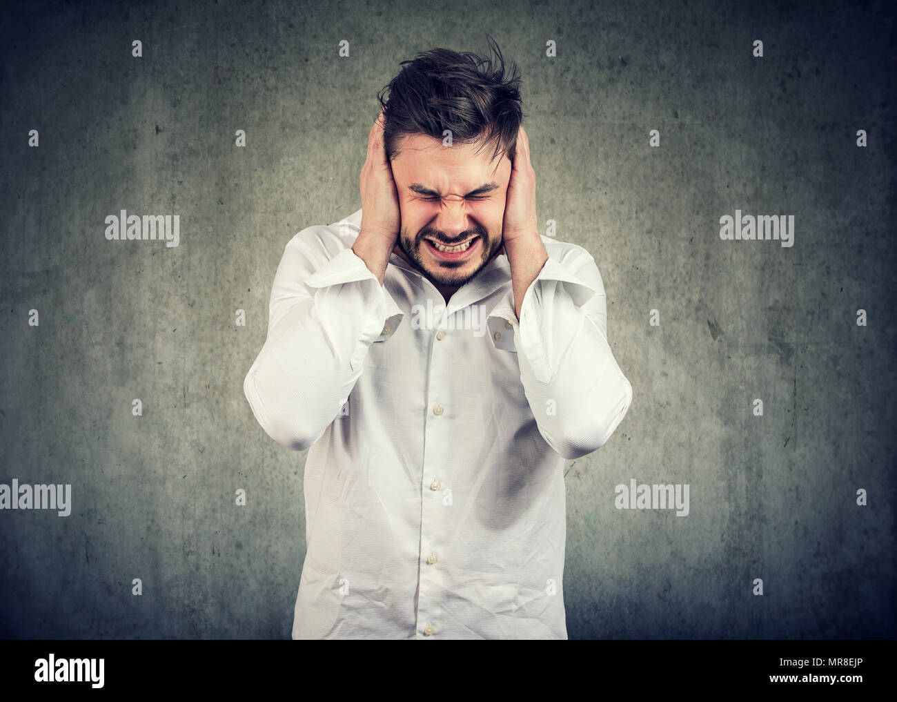 Disperata giovane uomo che copre le orecchie e accigliata a causa di stress e la pressione su sfondo grigio. Foto Stock