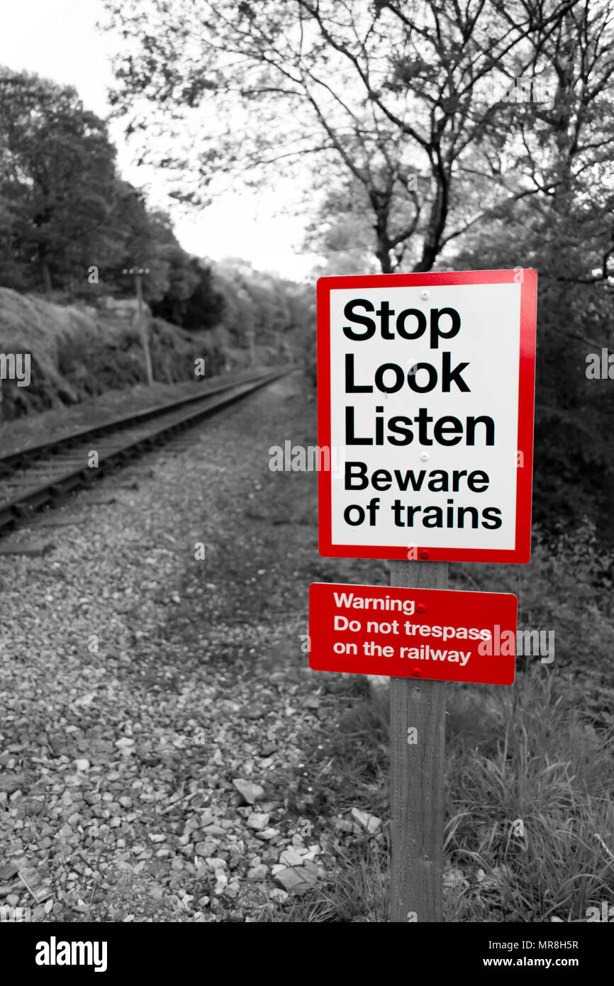 Stazione ferroviaria segno di avvertimento in bianco e nero con rosso lettura signpost 'Stop guardare ascoltare attenti di treni' con una linea ferroviaria in esecuzione fuori nella distanza. Foto Stock
