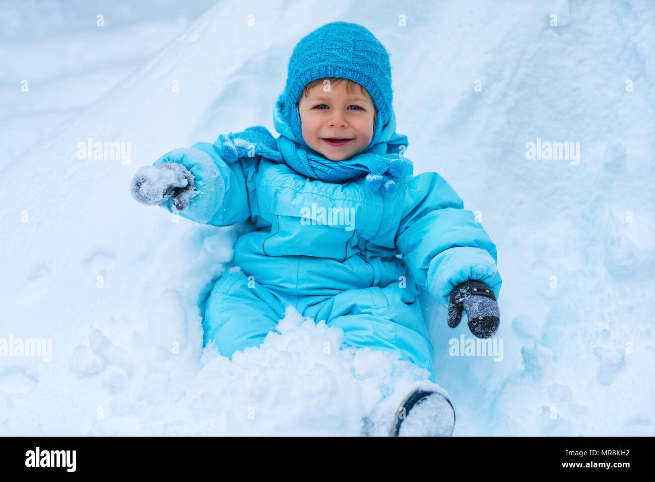 Due anni di bambino in tuta blu seduti sulla neve e sorridente Foto Stock