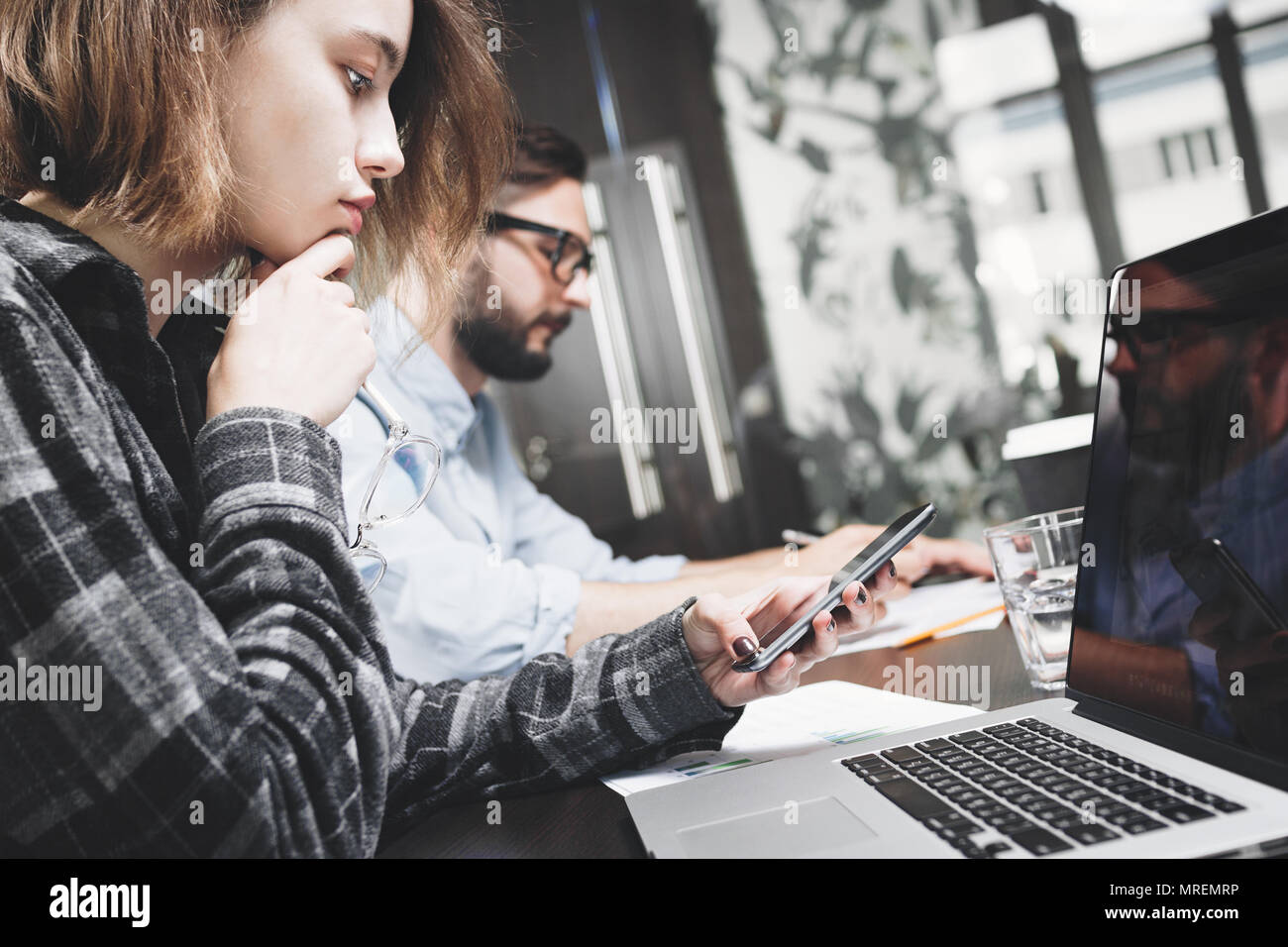 Giovane imprenditrice con gli occhiali e lo smartphone in mano funziona con computer portatile. Lavorare con dispositivi digitali in moderno spazio loft Foto Stock