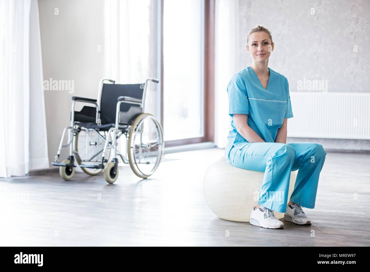 Femmina giovane fisioterapista seduti sulla palla Svizzera con la sedia a rotelle in background. Foto Stock