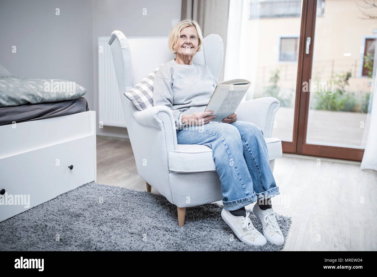 Senior donna seduta in poltrona con la rivista in casa di cura. Foto Stock