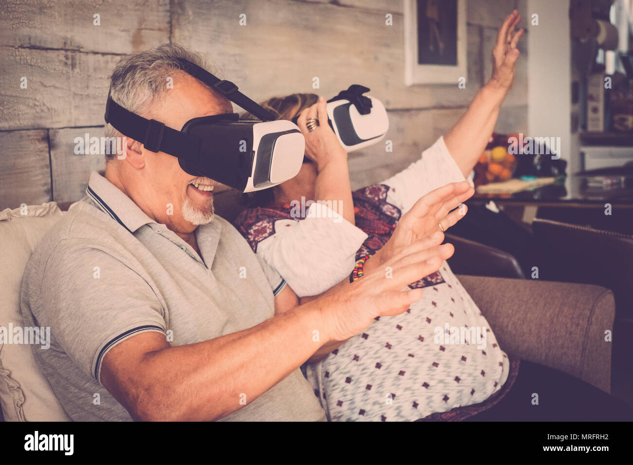 Senior l uomo e la donna con gli occhiali di protezione auricolare di realtà virtuale occhiali di giocare e divertirsi seduti sul divano di casa. Filtro a caldo e la vita insieme c Foto Stock