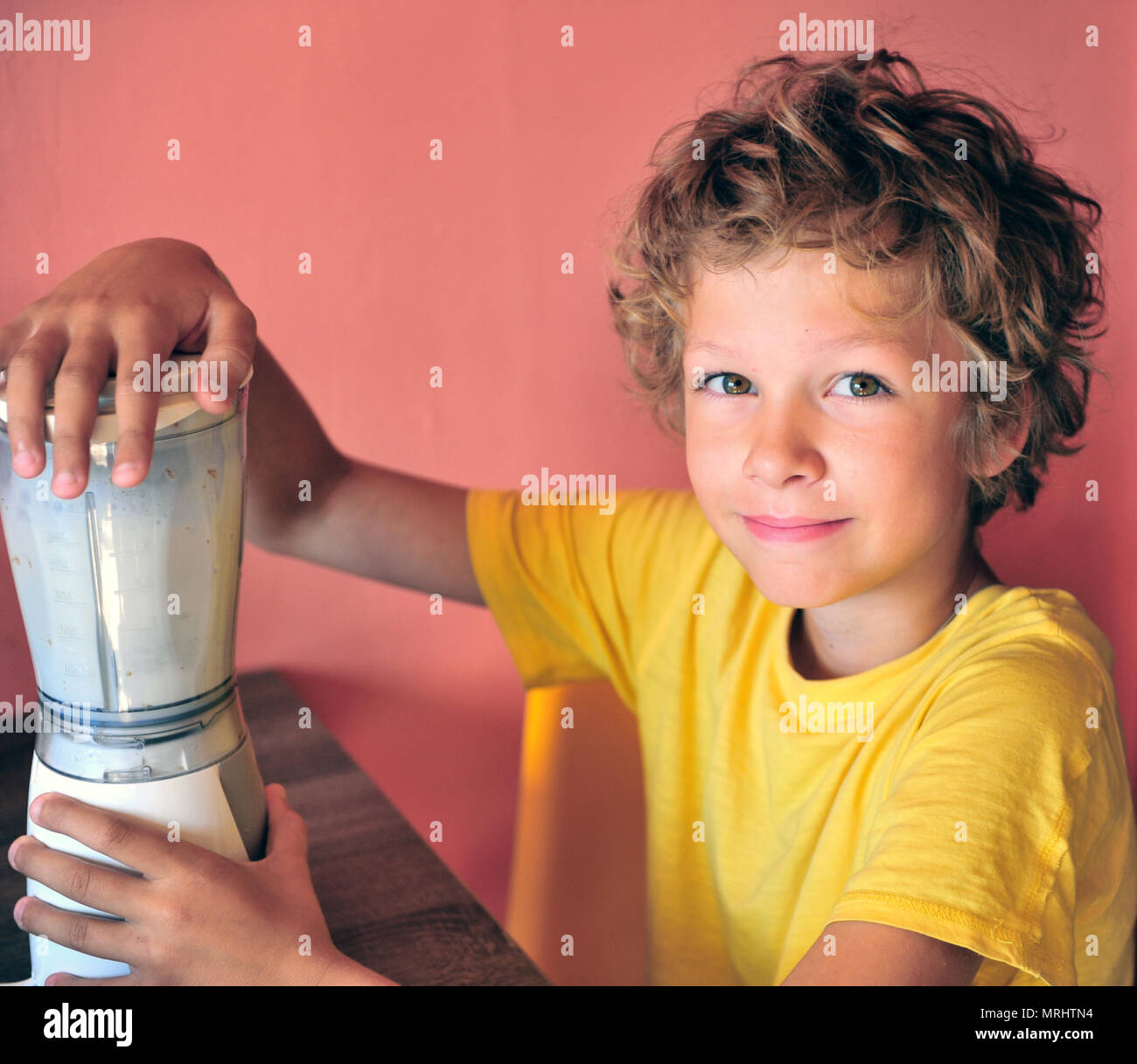 Portrair di un ragazzo felice rendendo milk shake per se stesso seduto alla cucina, adorabili caucasian capretto con shaggy capelli Foto Stock