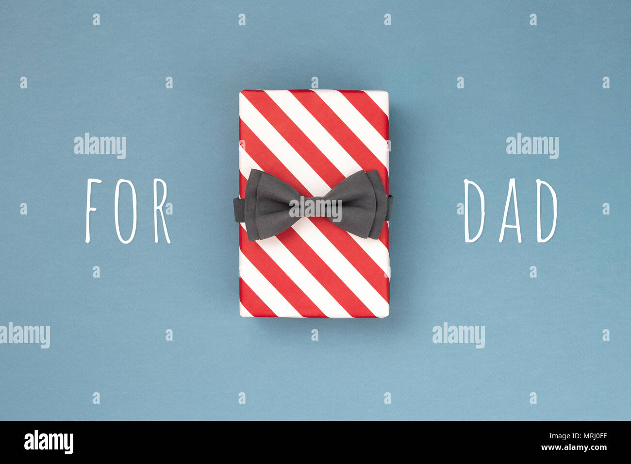 Una scatola regalo fasciata in rosso a strisce di carta e legato con il grigio bow tie su blu-grigio. L'idea di design regalo per un papà. Concetto di vacanza. Foto Stock