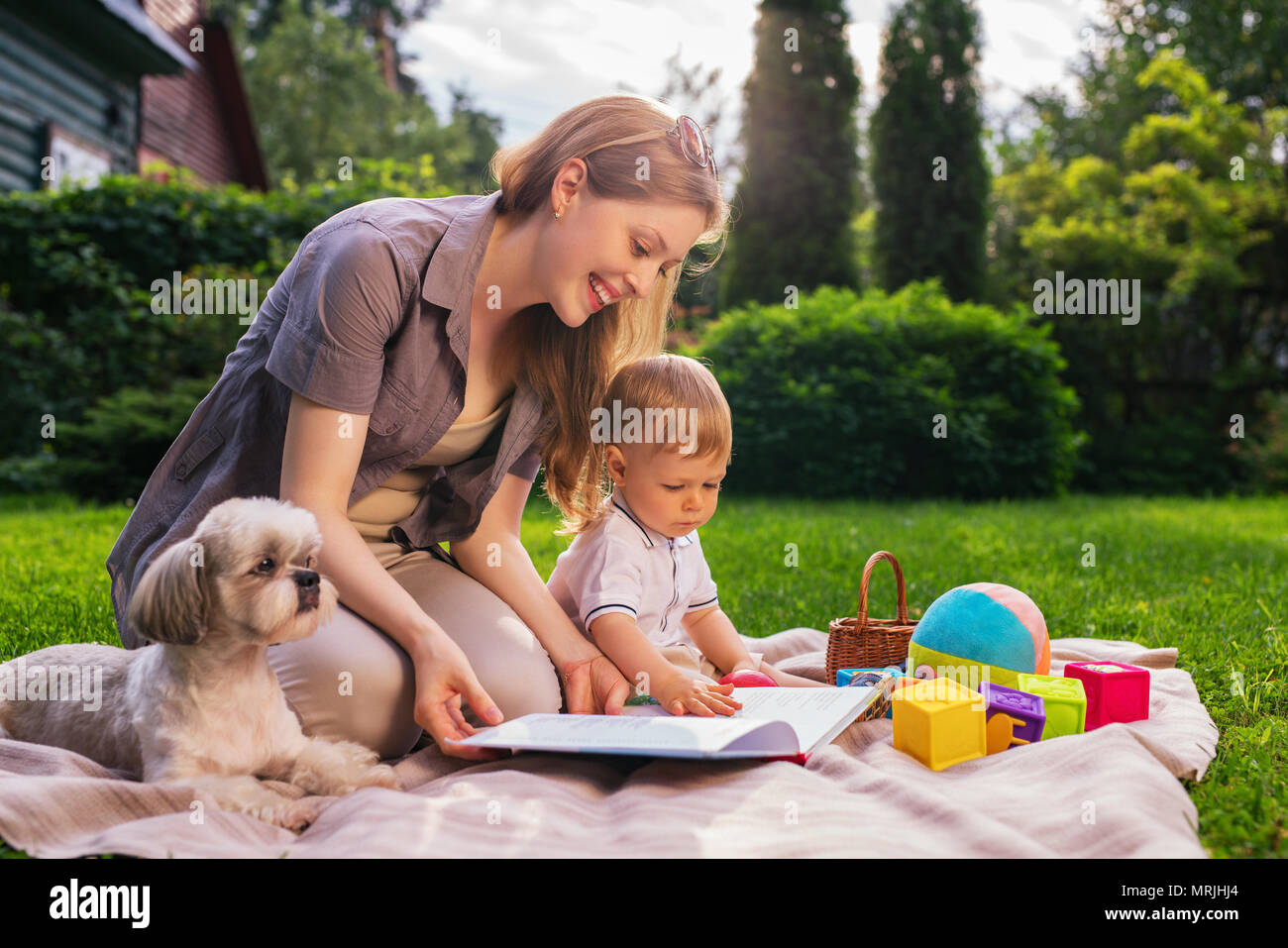 Giovane madre con un anno di bambino e dog sitter nel parco e libro di lettura Foto Stock