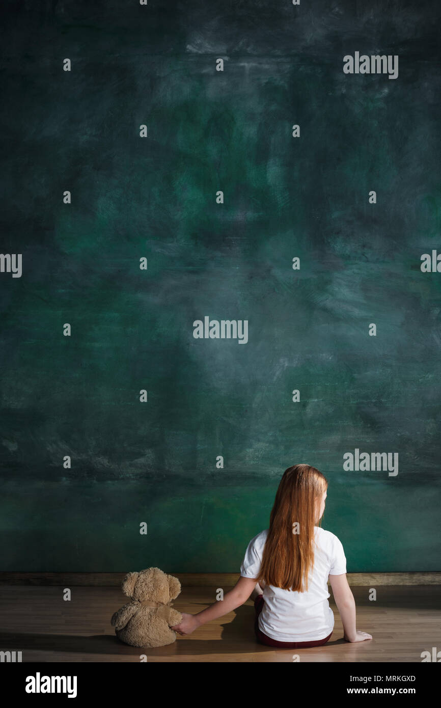 Bambina con orsacchiotto seduto sul pavimento nella stanza vuota. Concetto di autismo Foto Stock