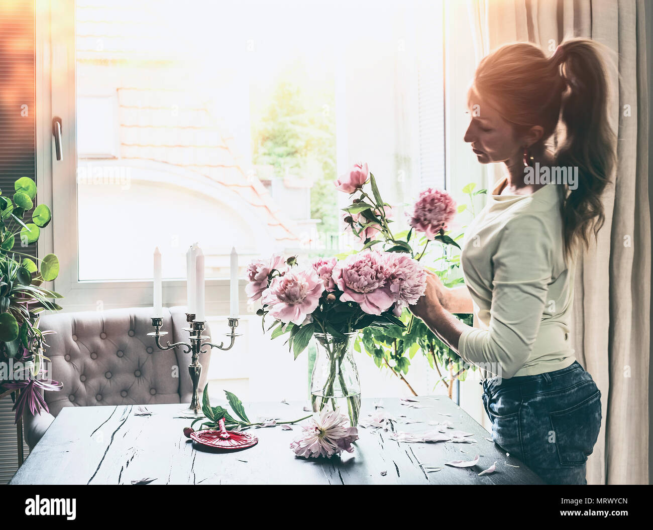 Bella donna moderna con pony tail hair disponendo le peonie mazzetto in vaso di vetro sul tavolo in vetro in salotto. Stile di vita. Casa felice Foto Stock