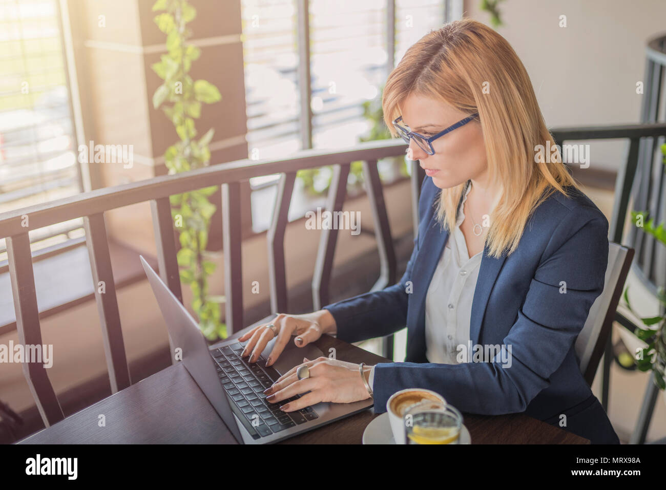 Bella donna d'affari con gli occhiali è l uso dei computer e di bere il caffè e un bicchiere di acqua in una caffetteria, ristorante. Affascinante ragazza utilizzando notebook t Foto Stock
